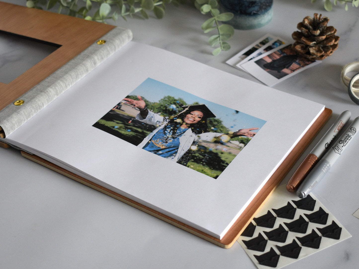 A graduation guest book lies open on a table with a 4x6 photograph of the graduate in their cap and gown. Photo holders lie next to the guest book for posting photo booth strips, polaroid pictures, and scrapbooking material.