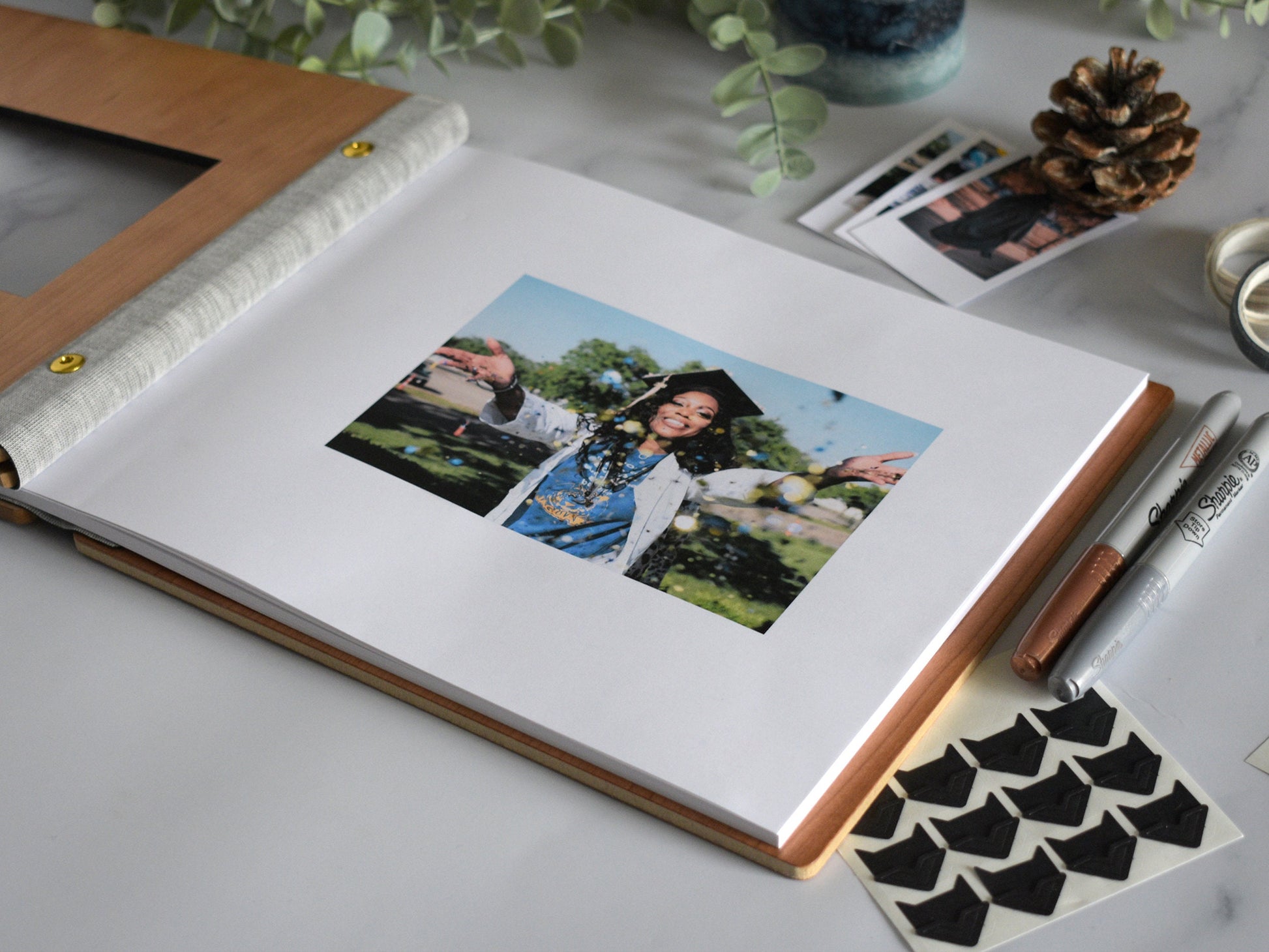 A graduation guest book lies open on a table with a 4x6 photograph of the graduate in their cap and gown. Photo holders lie next to the guest book for posting photo booth strips, polaroid pictures, and scrapbooking material.