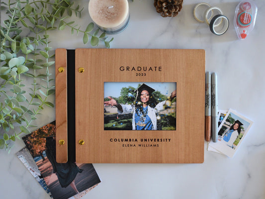 A graduation photo album lies on a table with polaroid photos of the graduate, washi tape, and double sided tape. This book serves as a graduation party decoration for guests to sign and post polaroids inside. It also makes the perfect grad gift.