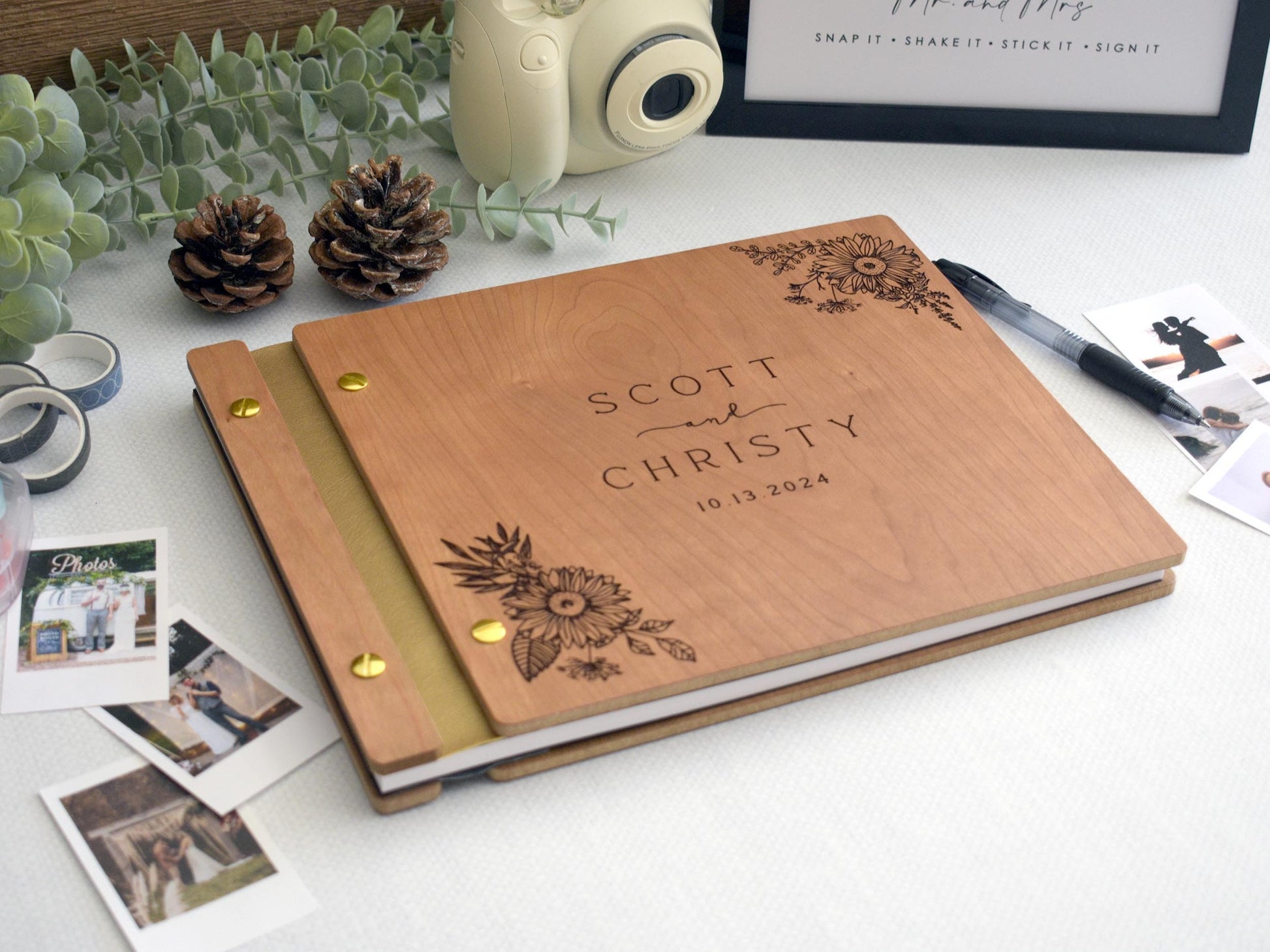 Custom cherry wood wedding guest book with an engraved floral sunflower design. Gold screws and gold leather binding. Surrounded by Polaroid photos, a pen, and photo tape, perfect for capturing special memories at weddings and significant events