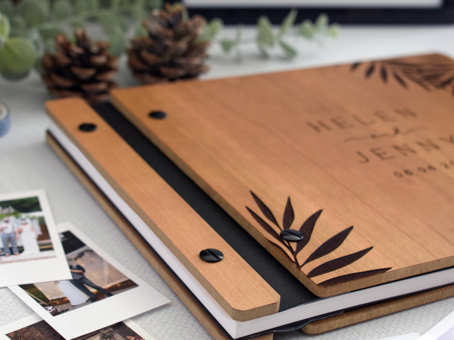 Custom wooden wedding guest book with tropical palm leaf design, black screws, and black binding. Surrounded by Polaroid photos, a pen, and photo tape, perfect for capturing memories at weddings, engagements, or anniversaries. Personalized keepsakes.