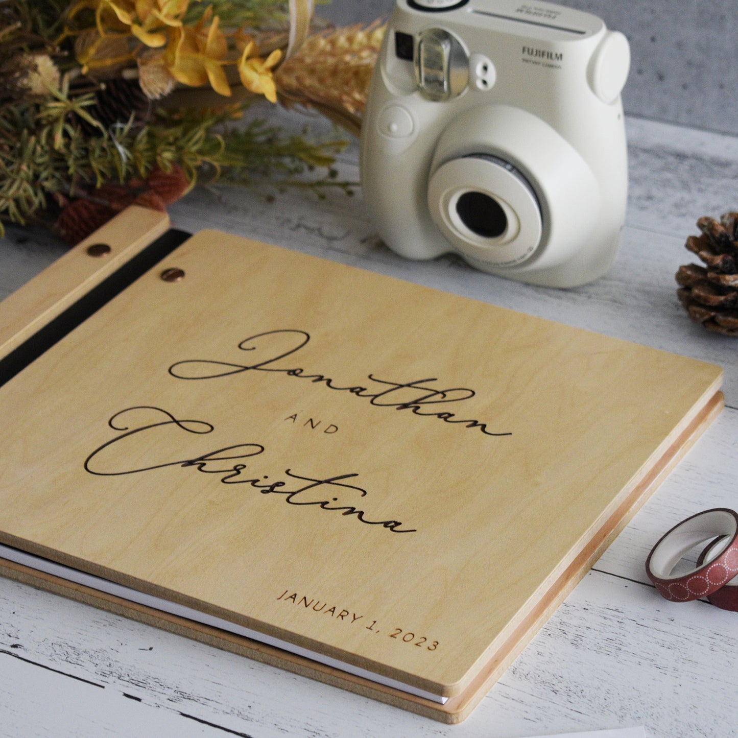 An 8.5x11" guest book lies on a table. Made of maple wood, black vegan leather binding, and copper hardware. The front cover reads “Jonathan and Christina, January 1, 2023.” 