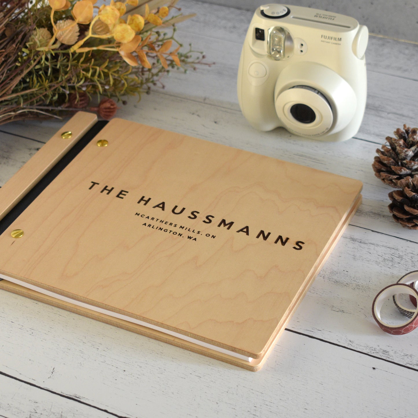 An 8.5x11" guest book lies on a table. Made of maple wood, black vegan leather binding, and gold hardware. The front cover includes an engraved design with personalized names.