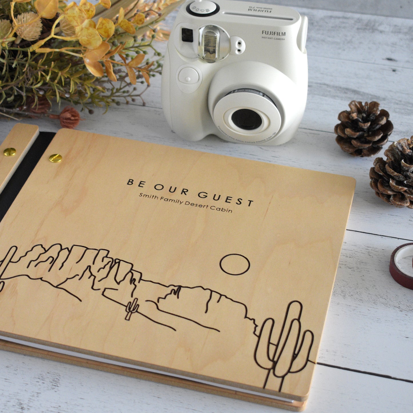 An 8.5x11" Airbnb guest book lies on a table. Made of maple wood, aqua vegan leather binding, and gold hardware. The front cover reads Be Our Guest, Smith Family Desert Cabin.