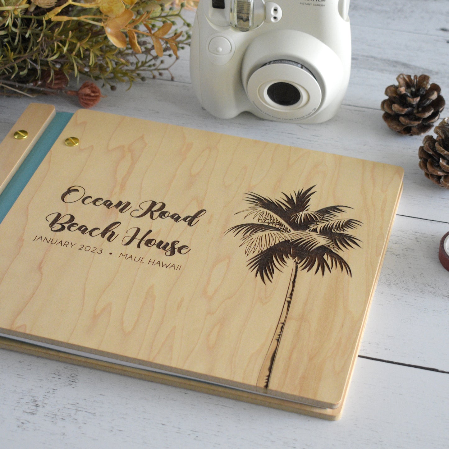 An 8.5x11" Airbnb guest book lies on a table. Made of maple wood, aqua vegan leather binding, and gold hardware. The front cover reads Ocean Road Beach House, January 2023, Maui, Hawaii.