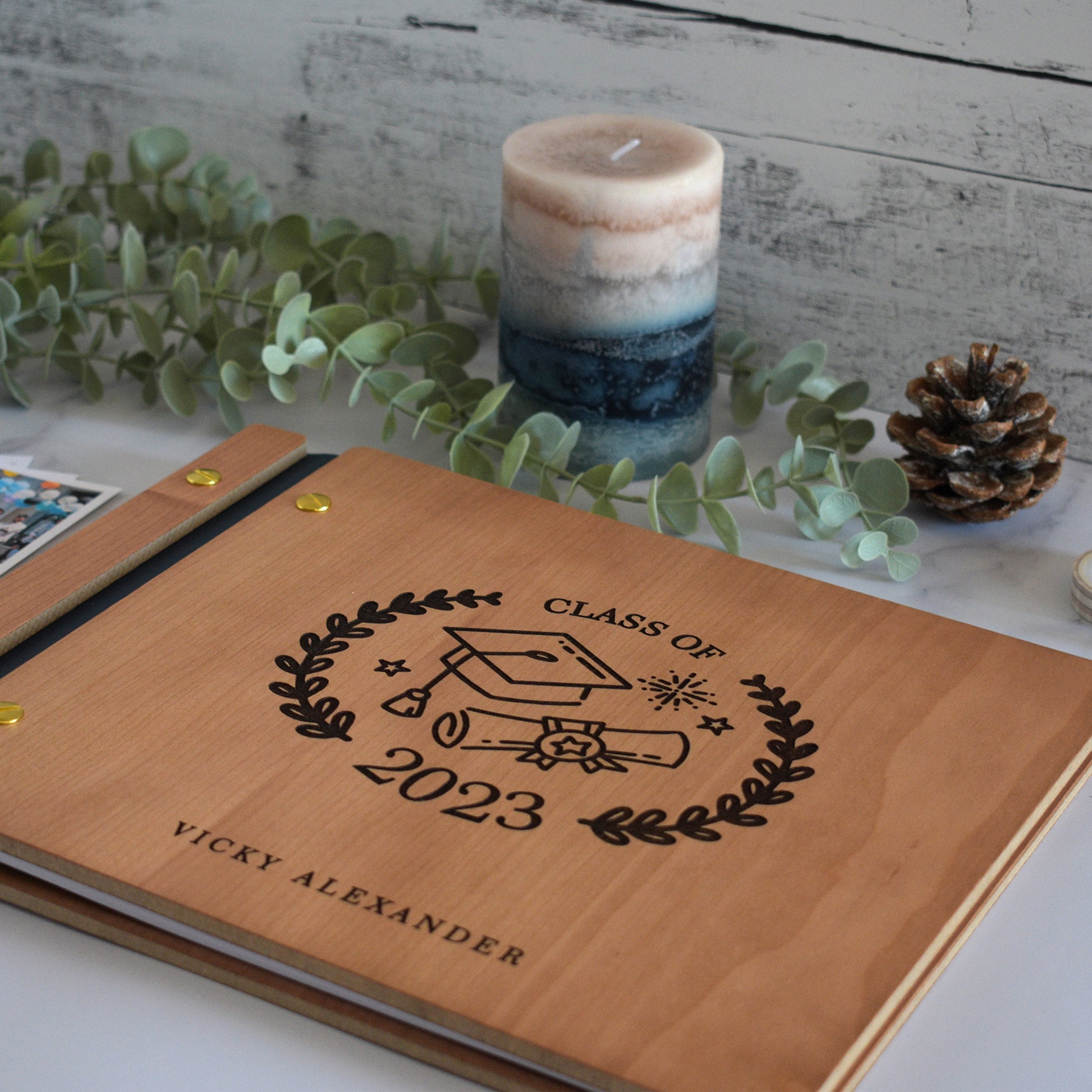 An 8.5x11" sized guest book lies on a table with greenery and a candle. The guest book includes a personalized engraving that reads Graduate 2023 along with the University name and Student name. The guest book also includes a cut out frame with a photo of the graduate.