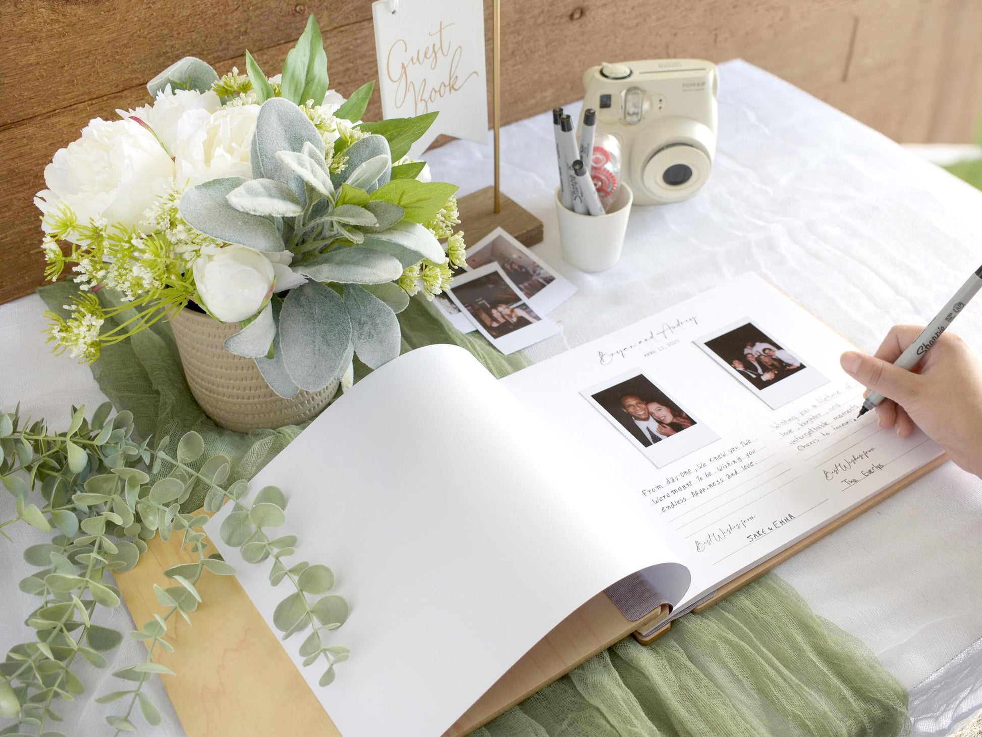 a guest book on a table with a person's hand writing in it. The guest book is surrounded by a bouquet of flowers, a camera, and some pens.