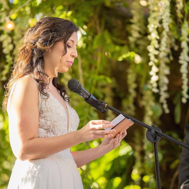A woman in a white wedding dress is speaking into a microphone at an outdoor ceremony, with a microphone stand in front of her.