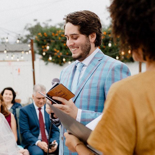 A man in a blue plaid suit is reading from a book during an outdoor ceremony, with other people in the background.