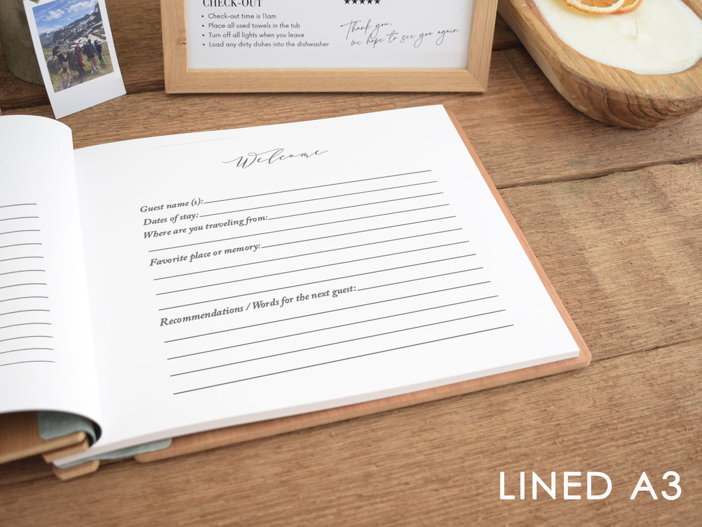 a lined guest book with a wooden frame, placed on a wooden surface. The book appears to be open, with a blank page visible. There is also a wooden bowl and a framed picture in the background.