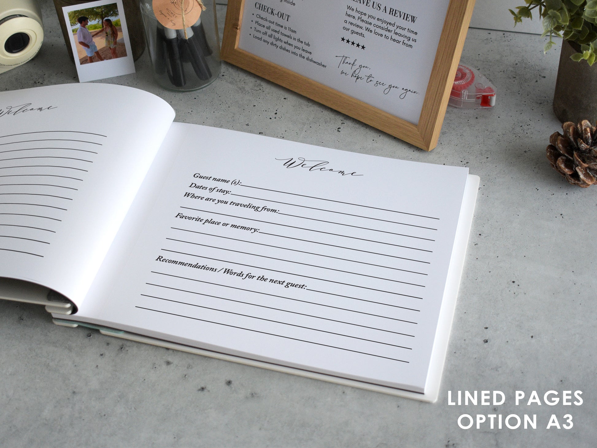 an open guest book with lined pages, placed on a table with various items such as a framed sign, a potted plant, and a camera.