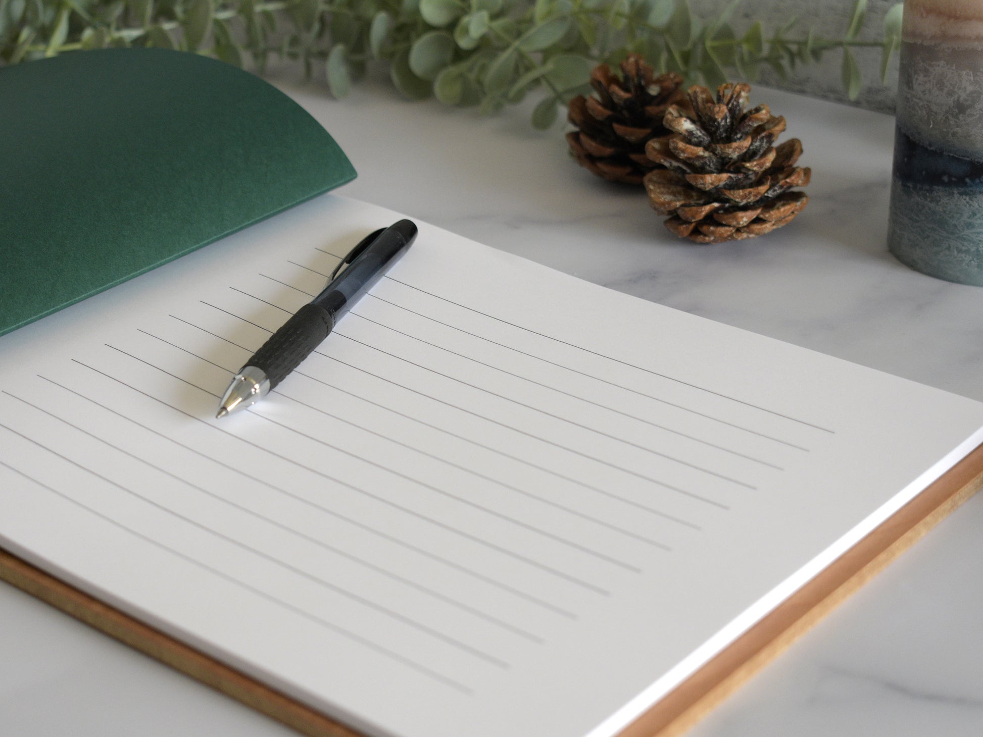 a lined notebook with a pen resting on top of it, placed on a marble surface. There are pine cones and a green folder nearby, creating a simple and organized workspace.