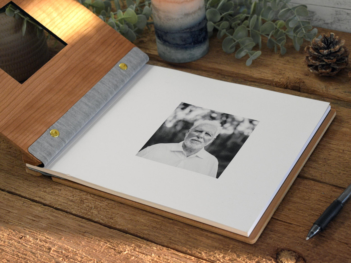 a wooden photo album with a black and white photograph of an older man on the cover, placed on a wooden surface. There is also a pen and a pine cone nearby.