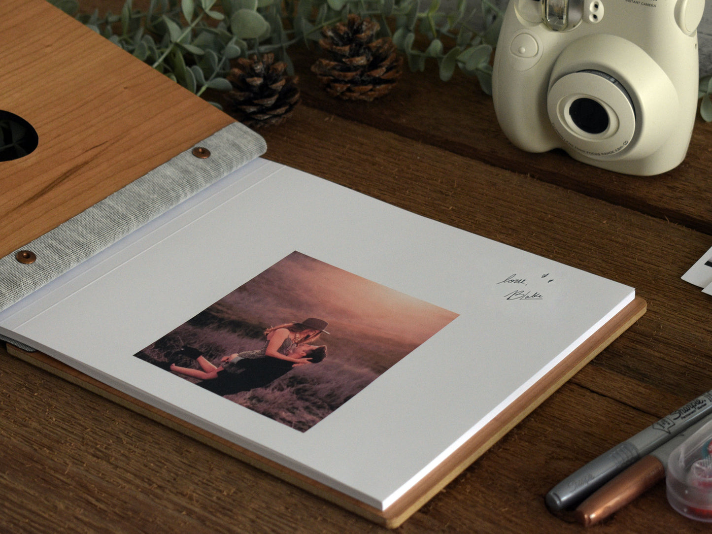 a wooden desk with a white instant camera, a photo album, and some pine cones.