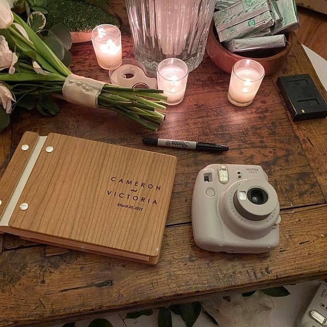 An 8.5X11 inch guest book with cherry wood, ivory leather binding, and silver hardware sits on a desk with a white polariod camera, pink bouquet of flowers, clear glass candles, and Fujifilm instax instant film packs.