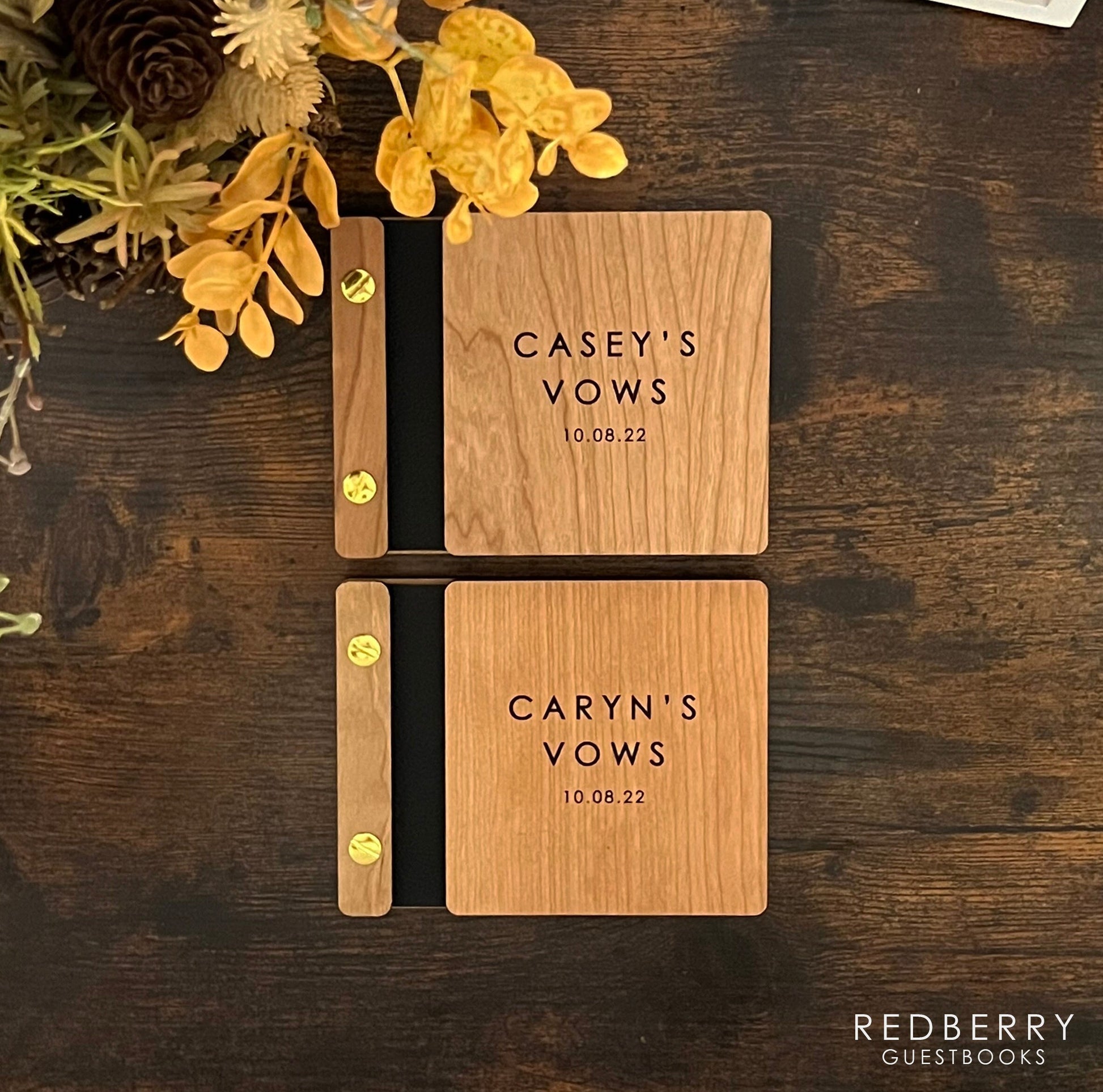 Two 5.25 X 4.25 matching vow books rest on a rustic table, made out of cherry wood, vegan leather, and gold aluminum hardware. The front cover of each book has custom engravings, that read Casey&#39;s Vows 10.08.22 and Caryn&#39;s Vows 10.08.22.