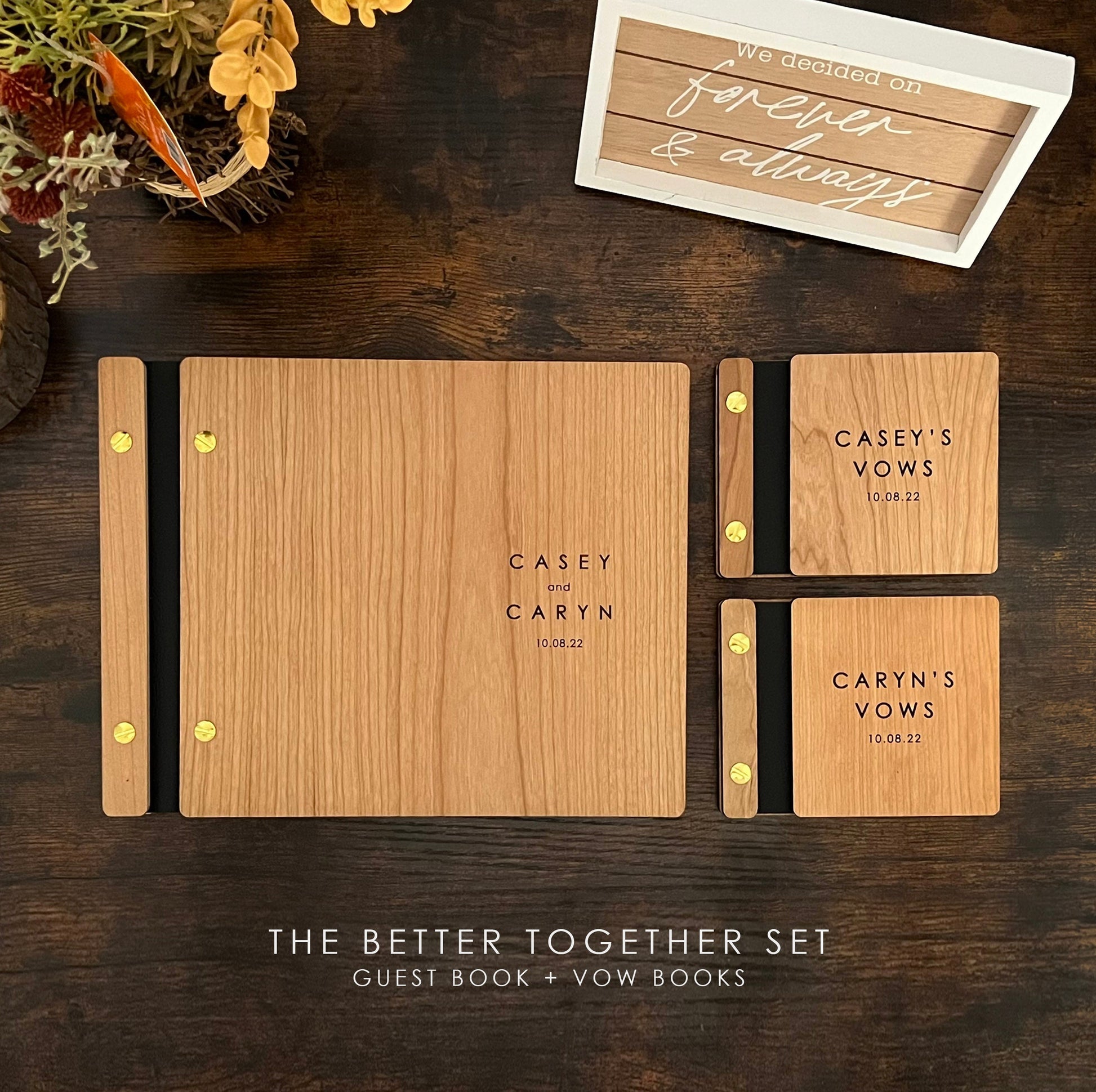 An 8.5X11 guest book sits at the center of a rustic table with two 5.25 X 4.25 vow books at its side. Made out of cherry wood, black binding, and gold screws. They read Casey and Caryn 10.08.22. with a sign and flowers in the background.