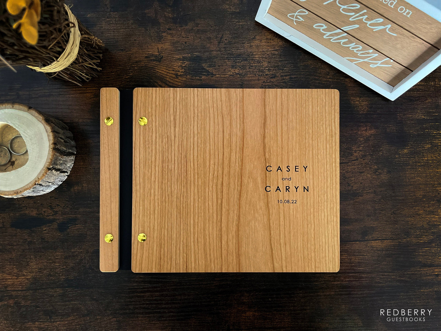 An 8.5X11 inch wedding guestbook sits on a rustic desk alongside a forever and always sign, flower bouquet, and a tree trunk-themed ring holder. Made out of cherry wood, black vegan leather, and gold screws, it reads Casey and Caryn 10.08.2022.