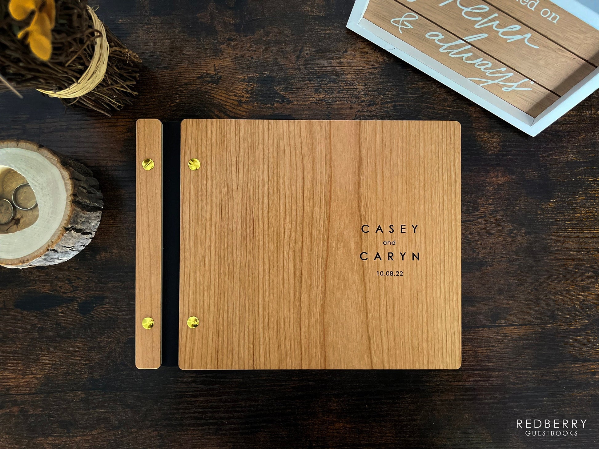 An 8.5X11 inch wedding guestbook sits on a rustic desk alongside a forever and always sign, flower bouquet, and a tree trunk-themed ring holder. Made out of cherry wood, black vegan leather, and gold screws, it reads Casey and Caryn 10.08.2022.