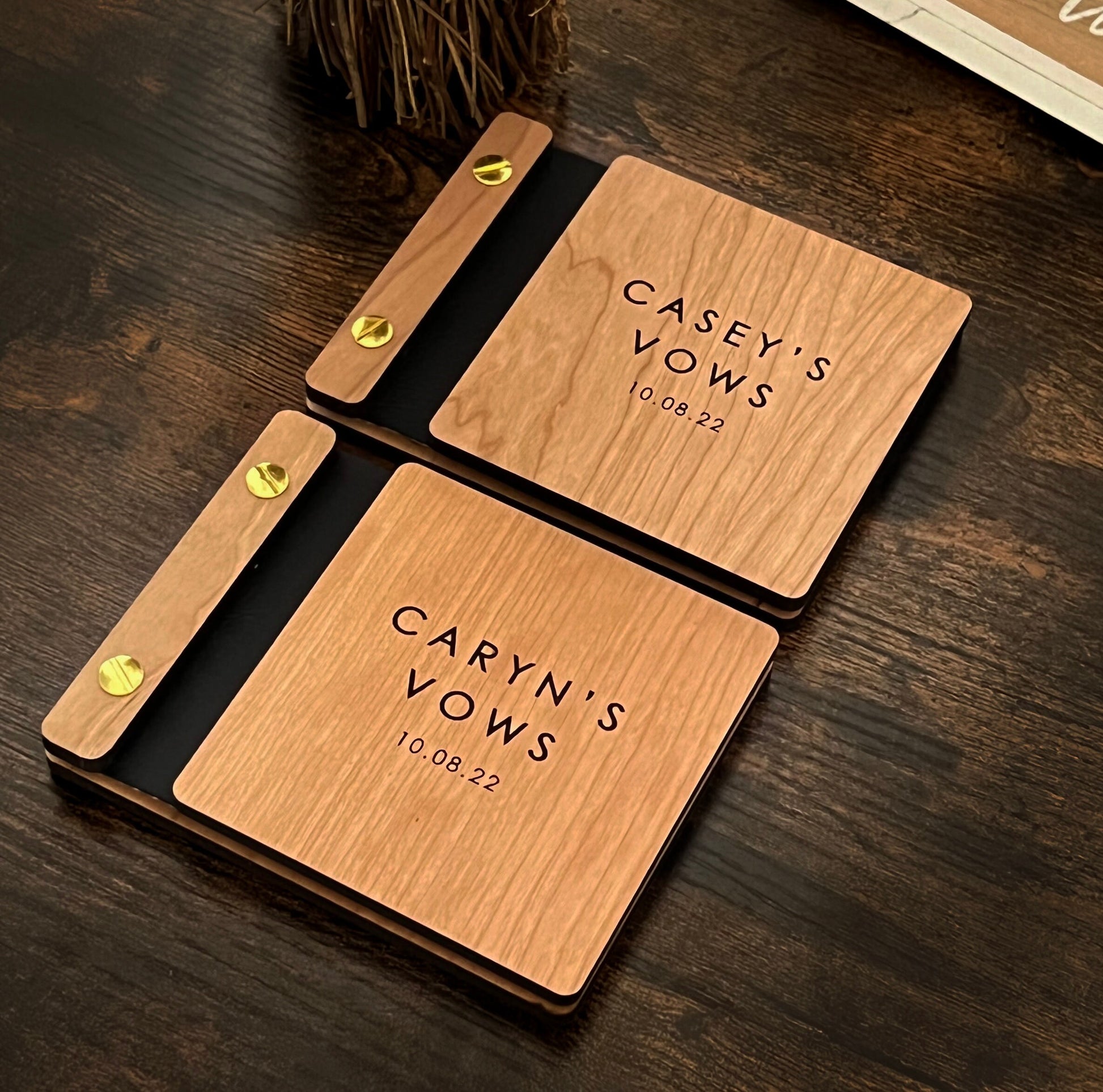 Two 5.25 X 4.25 matching vow books rest on a table at an angle, made out of cherry wood, vegan leather, and gold aluminum hardware. The front cover of each book has custom engravings, that read Casey&#39;s Vows 10.08.22 and Caryn&#39;s Vows 10.08.22.