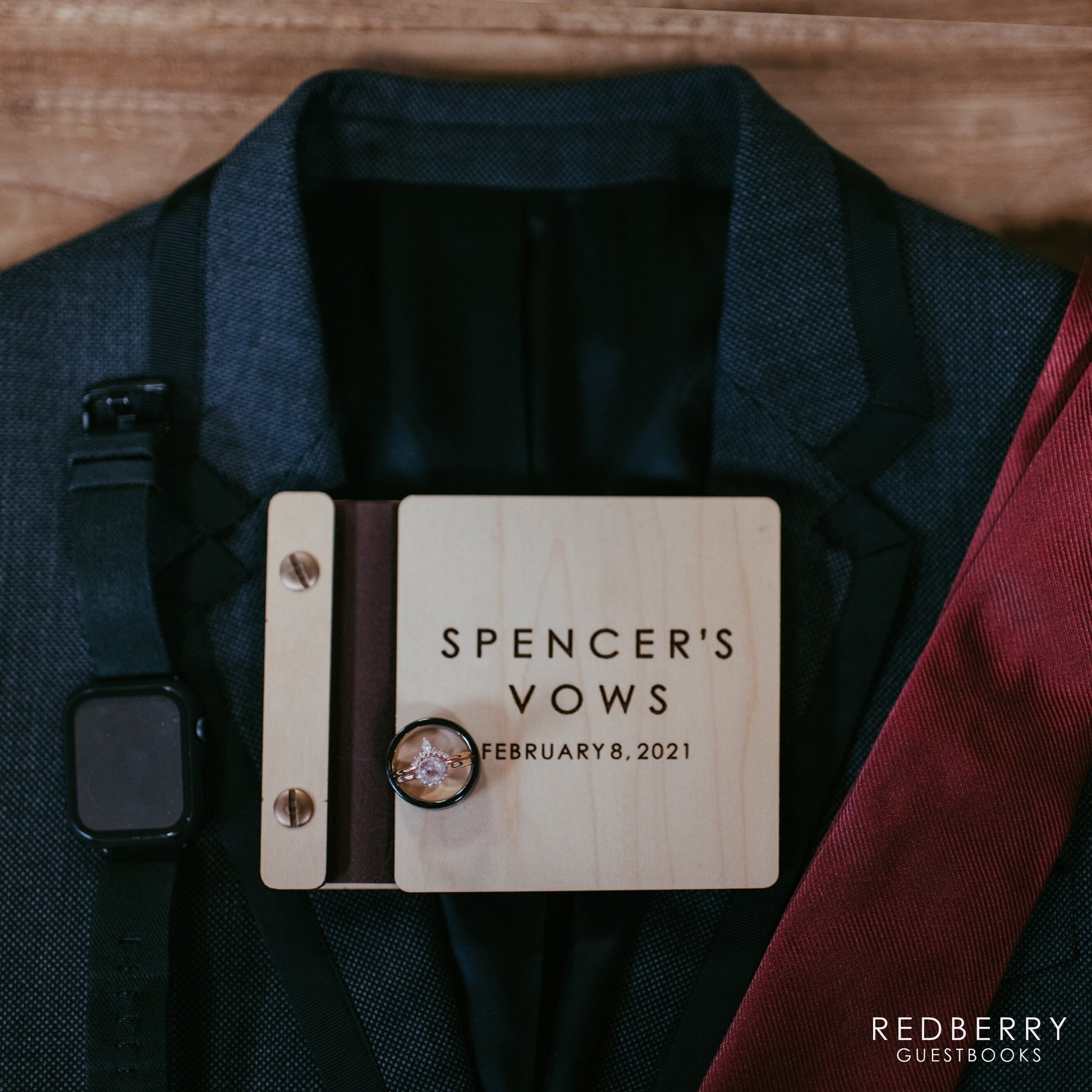 A 5.25 X 4.25 vow book made of maple wood, black binding, and silver screws sits on top of a blue suit, red tie, and black smartwatch with a black men&#39;s band and diamond wedding ring. The vow book reads Spencer&#39;s Vows, February 8, 2021.