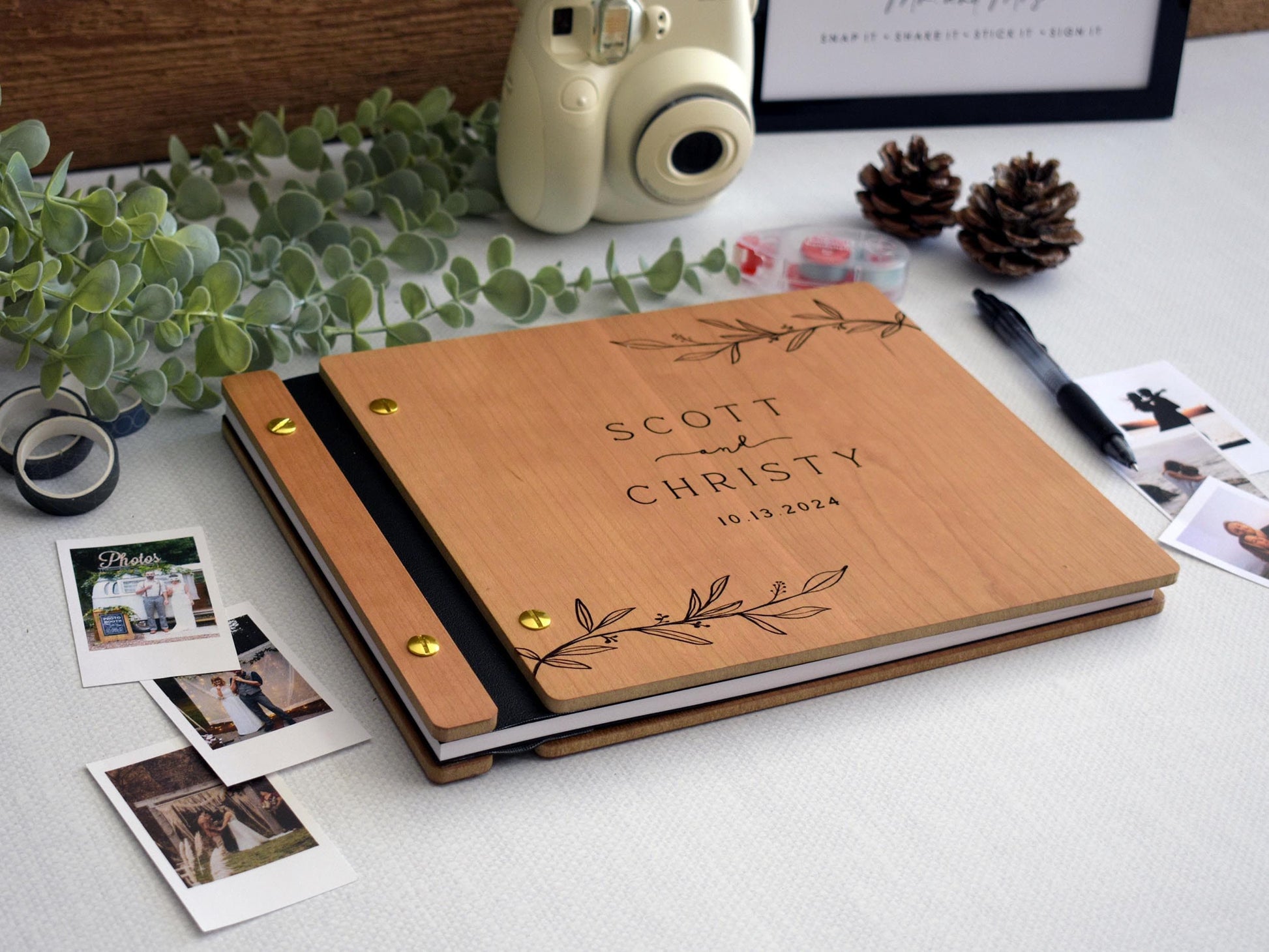 custom wedding guest book made of cherry wood, with gold screws and black binding. The cover displays an elegant engraved leaf motif. Surrounding the guest book are personalizing pages, black pen, Polaroid photos, photo tape, and washi tape.
