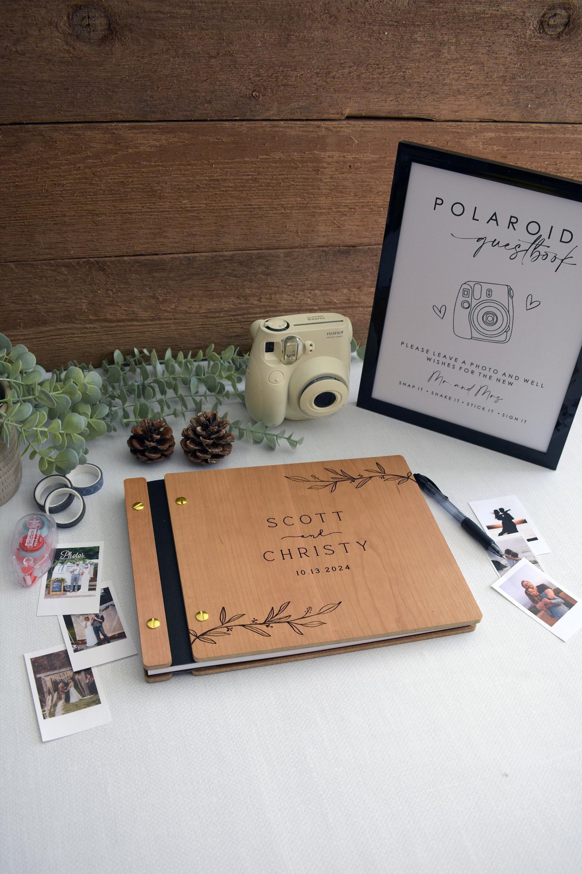 custom wedding guest book made of cherry wood, with gold screws and black binding. The cover displays an elegant engraved leaf motif. Surrounding the guest book are personalizing pages, black pen, Polaroid photos, photo tape, and washi tape.