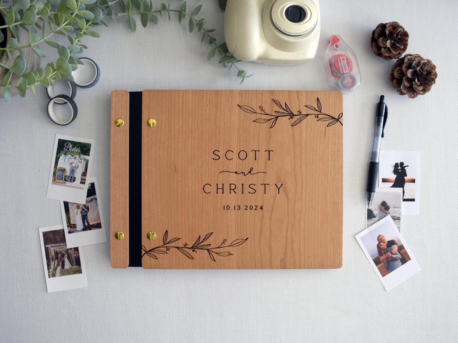 custom wedding guest book made of cherry wood, with gold screws and black binding. The cover displays an elegant engraved leaf motif. Surrounding the guest book are personalizing pages, black pen, Polaroid photos, photo tape, and washi tape.