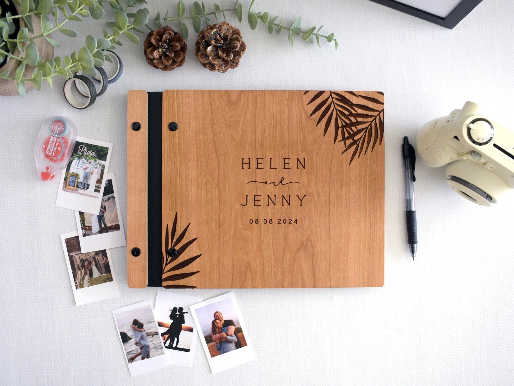 Custom wooden wedding guest book with tropical palm leaf design, black screws, and black binding. Surrounded by Polaroid photos, a pen, and photo tape, perfect for capturing memories at weddings, engagements, or anniversaries. Personalized keepsakes.