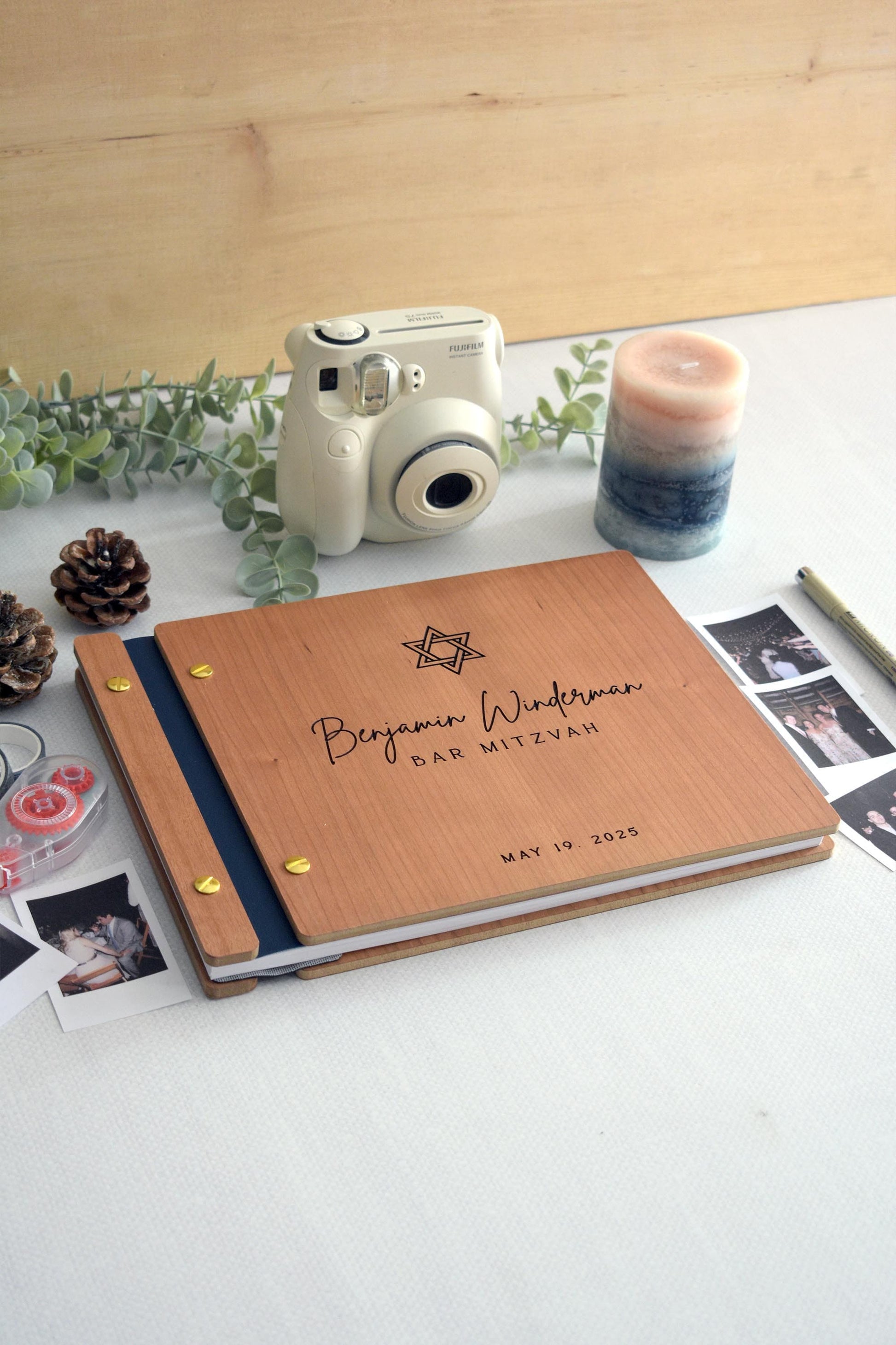 Custom Bar Mitzvah guest book featuring a Star of David design. The book is surrounded by polaroid photos, pinecones, a candle, a plant, and a pen, perfect for capturing memories. Polaroid camera and washi tape in the background. Fully customizable.