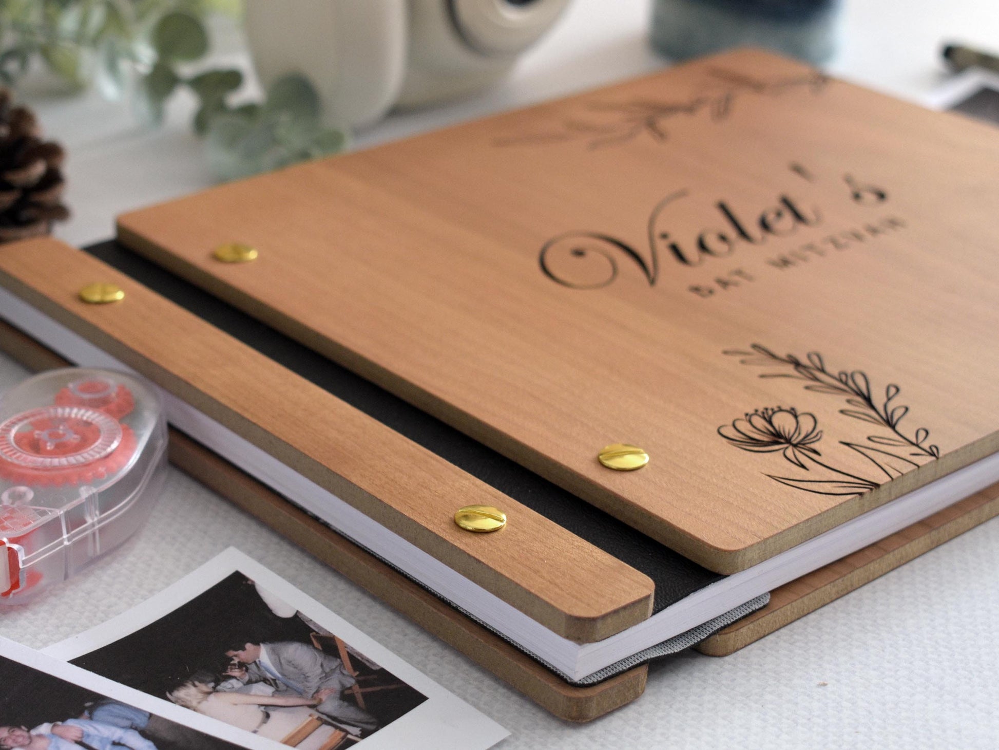 Wooden guest book with Bat Mitzvah engraved on the cover, decorated with elegant floral designs. Surrounding items include a Polaroid camera, photos, tape rolls, a candle, and a pen. Perfect for capturing special moments at a bat mitzvah celebration.