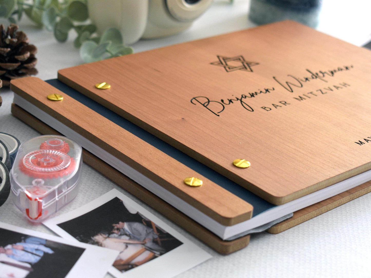 Close-up of a custom Bar Mitzvah guest book with a Star of David design and personalized text. The wooden cover is secured with gold screws and a navy blue spine. Surrounding items are polaroid photos, adhesive tape and pinecones. Memorable keepsake.