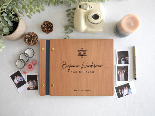 Custom Bar Mitzvah guest book featuring a Star of David design. The book is surrounded by polaroid photos, pinecones, a candle, a plant, and a pen, perfect for capturing memories. Polaroid camera and washi tape in the background. Fully customizable.