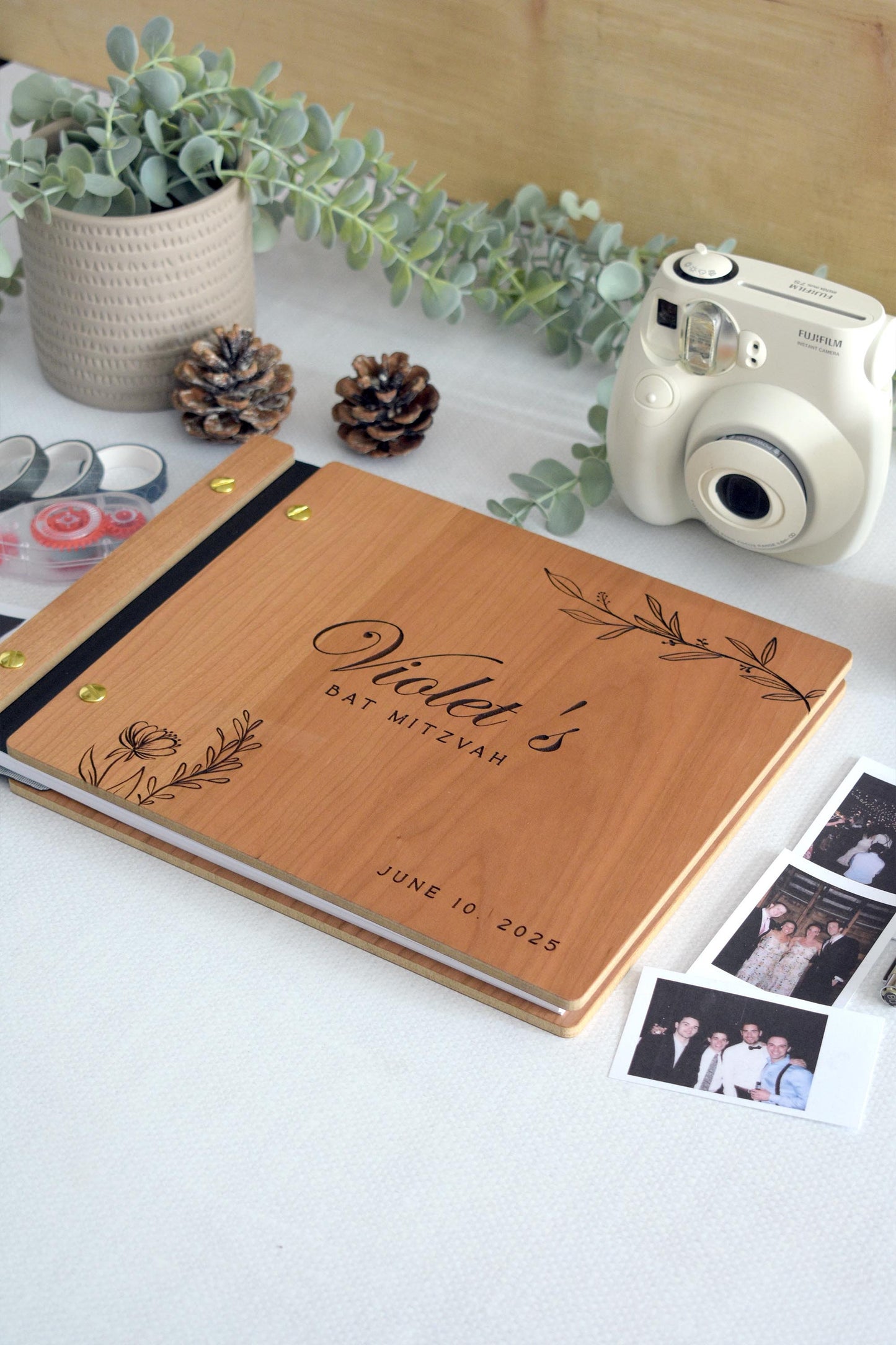 Wooden guest book with Bat Mitzvah engraved on the cover, decorated with elegant floral designs. Surrounding items include a Polaroid camera, photos, tape rolls, a candle, and a pen. Perfect for capturing special moments at a bat mitzvah celebration.