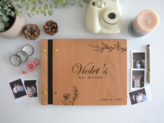Wooden guest book with Bat Mitzvah engraved on the cover, decorated with elegant floral designs. Surrounding items include a Polaroid camera, photos, tape rolls, a candle, and a pen. Perfect for capturing special moments at a bat mitzvah celebration.