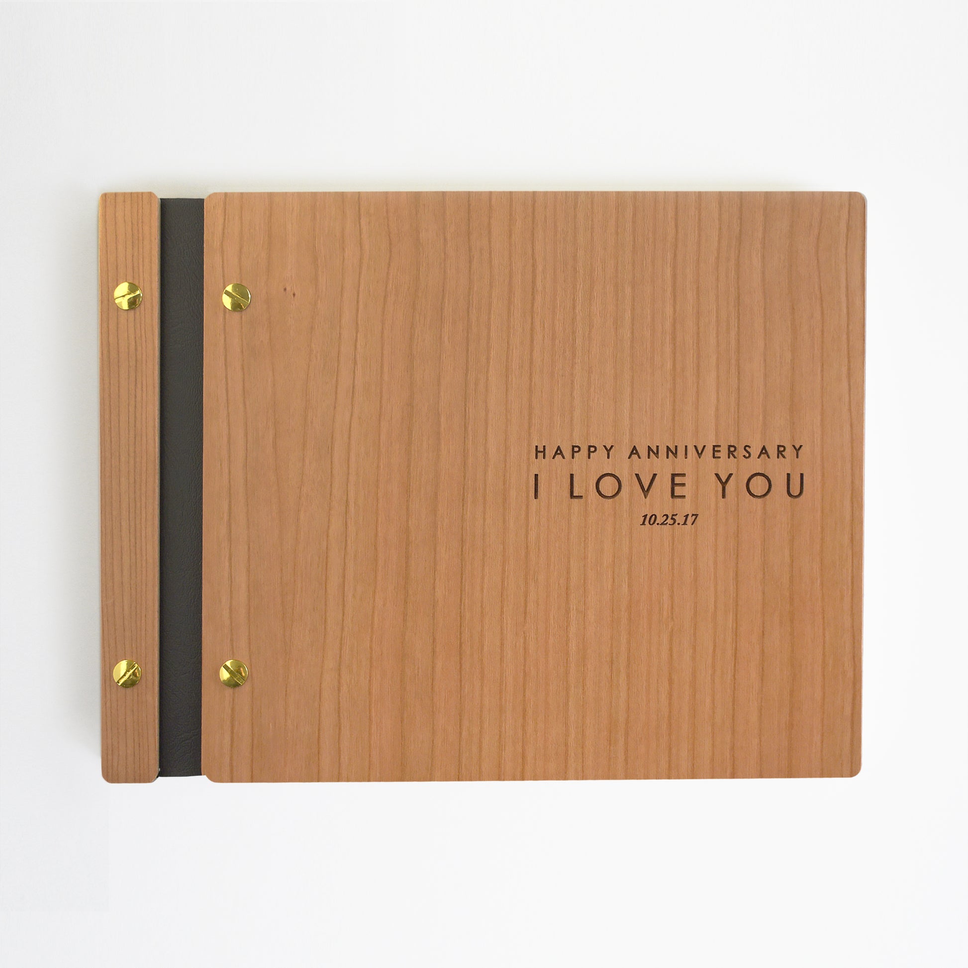 An 8.5x11" guest book lies on a table. Made of cherry wood, black vegan leather binding, and gold hardware. The front cover reads “Happy Anniversary, I love you. 10.25.17.”