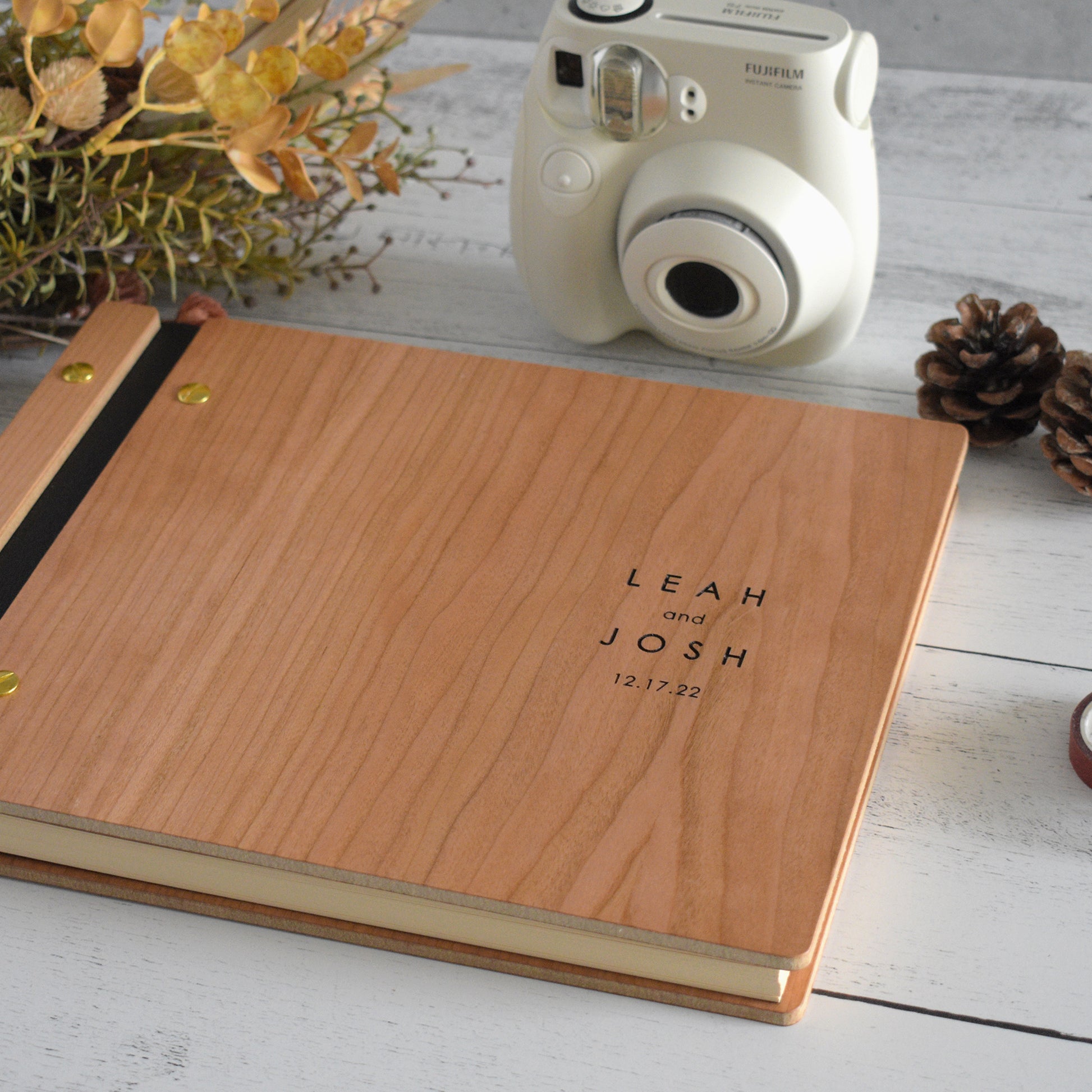 An 8.5x11" guest book lies on a table. Made of cherry wood, black vegan leather binding, and gold hardware. The front cover reads "Leah and Josh, 12.17.22."