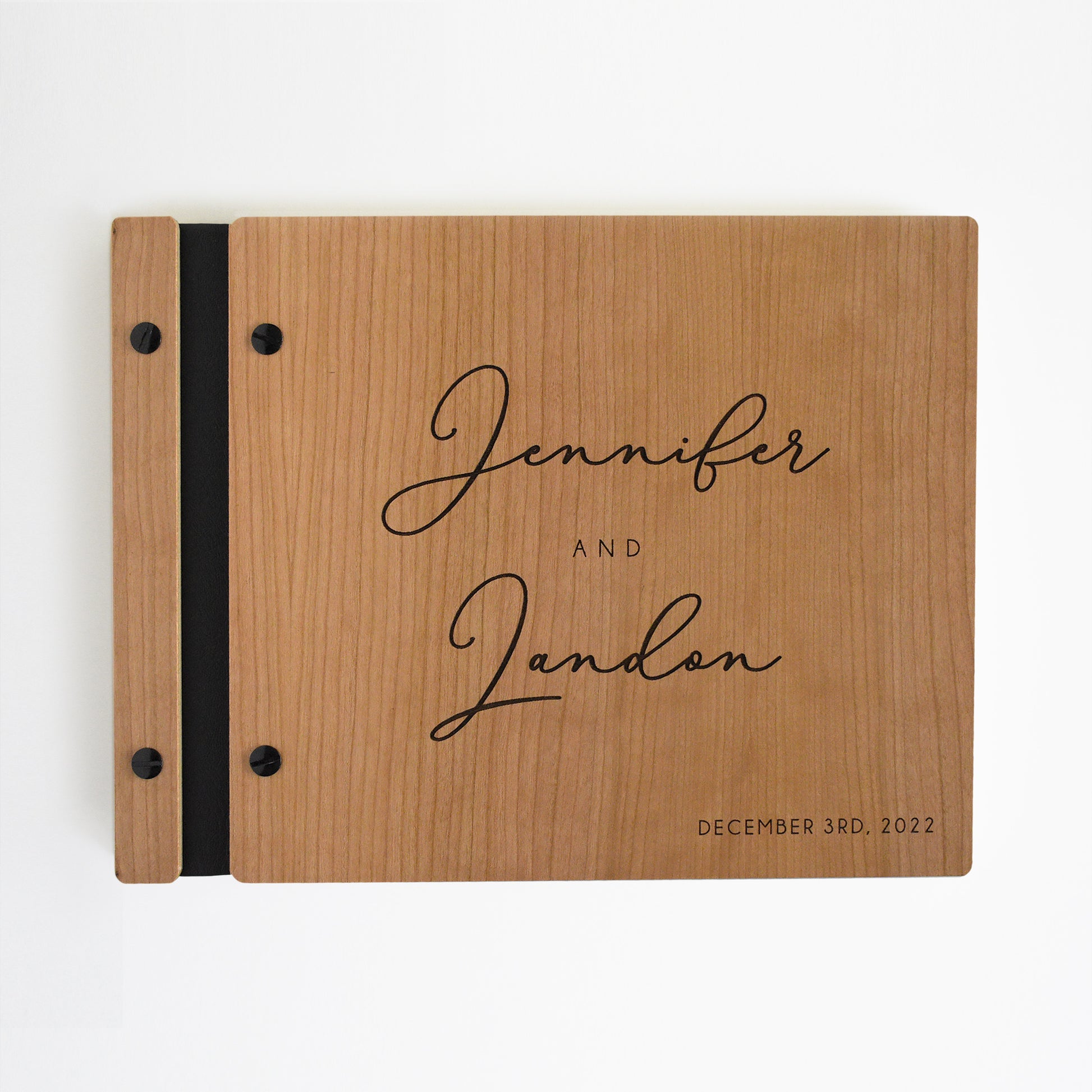 An 8.5x11" guest book lies on a table. Made of cherry wood, black vegan leather binding, and copper hardware. The front cover reads “Jonathan and Christina, January 1, 2023.” 