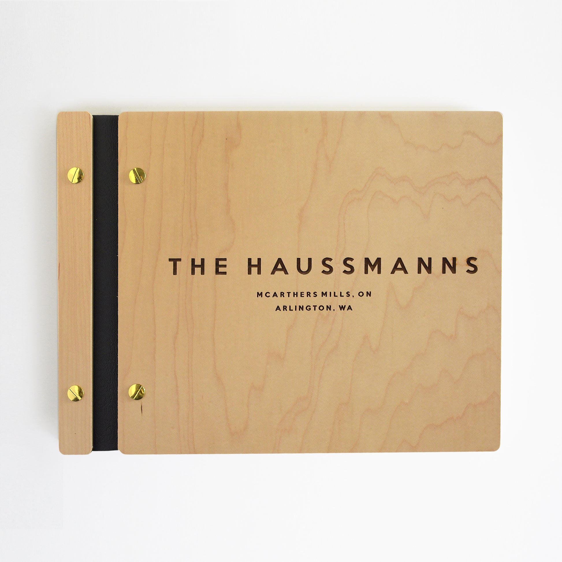 An 8.5x11" guest book lies on a table. Made of maple wood, black vegan leather binding, and gold hardware. The front cover includes an engraved design with personalized names.