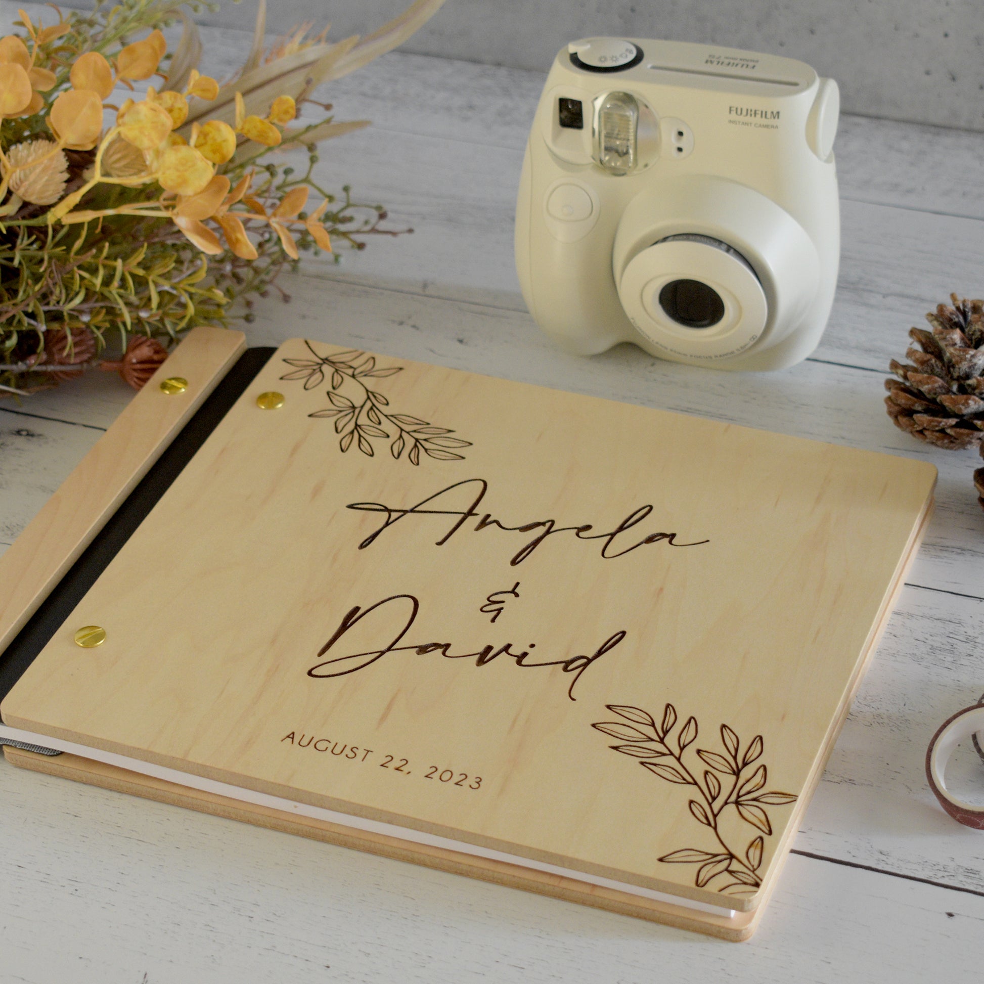 An 8.5x11" guest book lies on a table. Made of maple wood, black vegan leather binding, and gold hardware. The front cover has engravings of florals and reads Angela & David, August 22, 2023.
