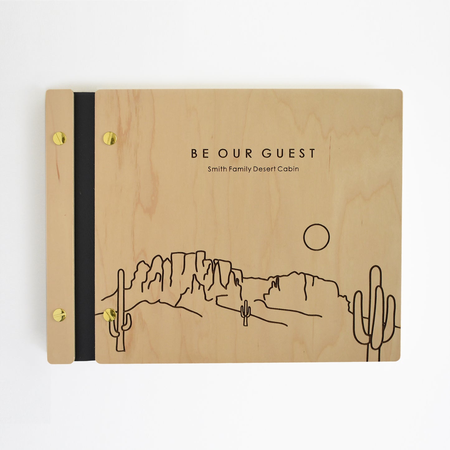 An 8.5x11" Airbnb guest book lies on a table. Made of maple wood, aqua vegan leather binding, and gold hardware. The front cover reads Be Our Guest, Smith Family Desert Cabin.