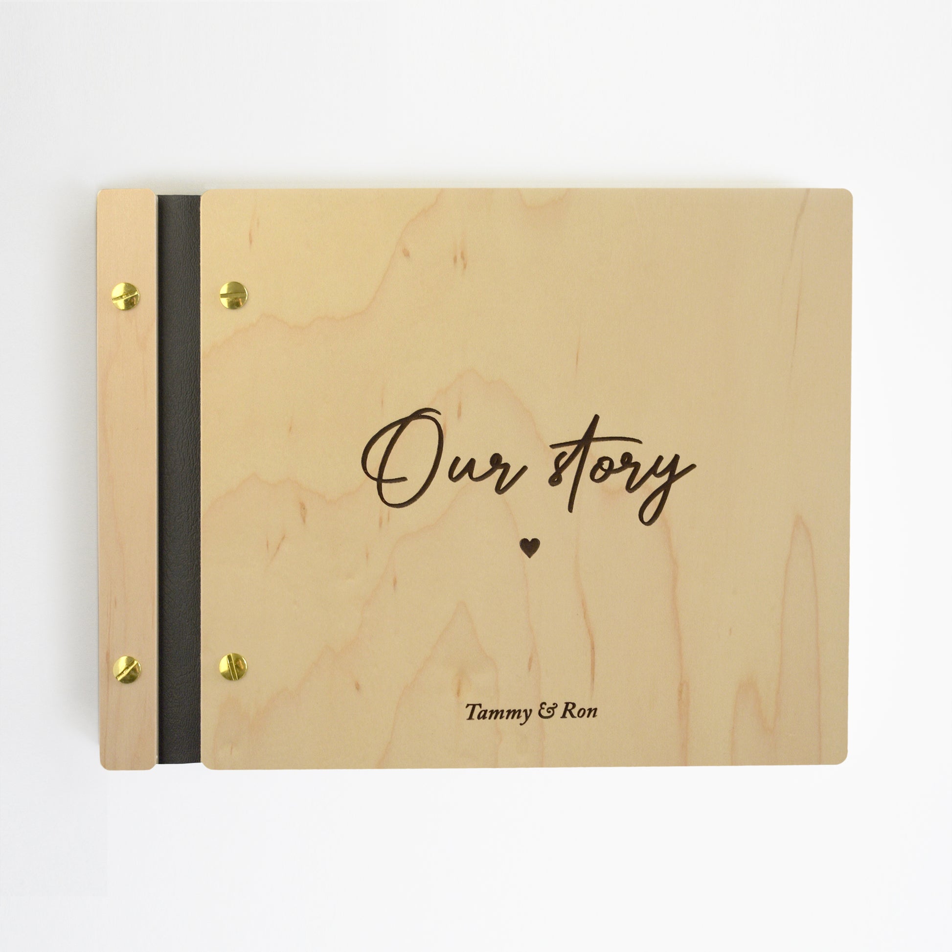 An 8.5x11" guest book lies on a table. Made of maple wood, black vegan leather binding, and gold hardware. The front cover reads “Our Story, Ron & Tammy.”