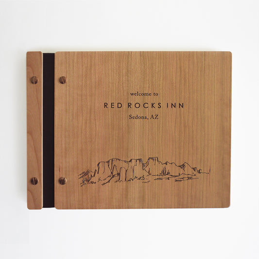 An 8.5x11" Airbnb guest book lies on a table. Made of cherry wood, black vegan leather binding, and copper hardware. The front cover reads Welcome to Red Rocks Inn, Sedona, AZ.