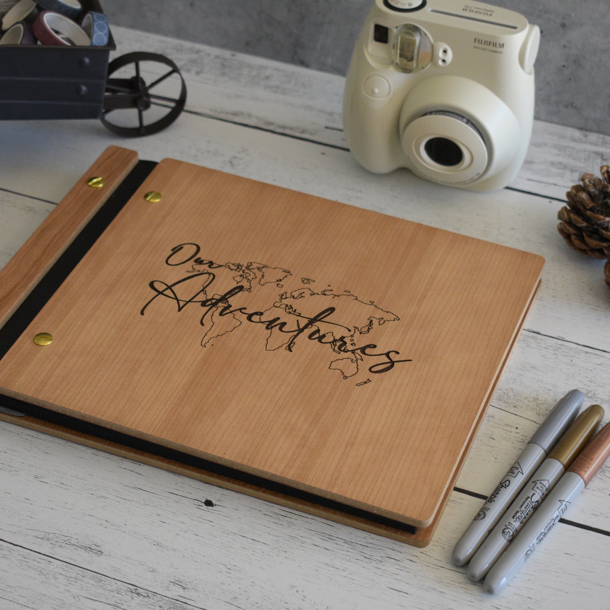 An 8.5x11 sized photo album with a white background sits on a desk. Made out of cherry wood, black vegan leather, and gold hardware, the book reads Our Adventures and has an engraving of a world map.