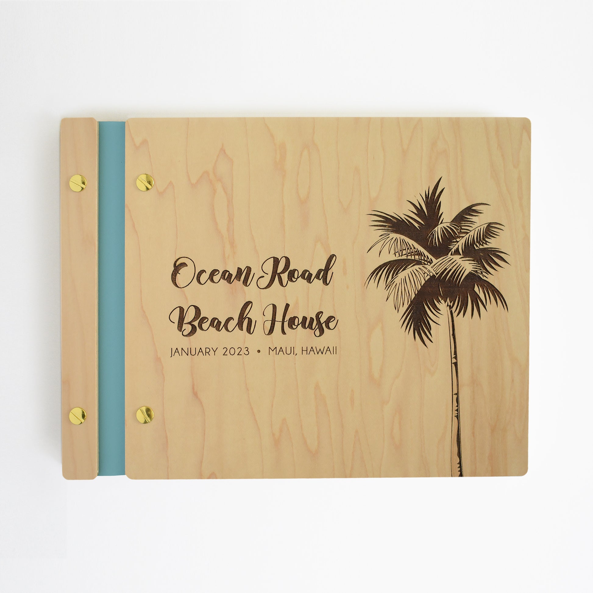 An 8.5x11" Airbnb guest book lies on a table. Made of maple wood, aqua vegan leather binding, and gold hardware. The front cover reads Ocean Road Beach House, January 2023, Maui, Hawaii.