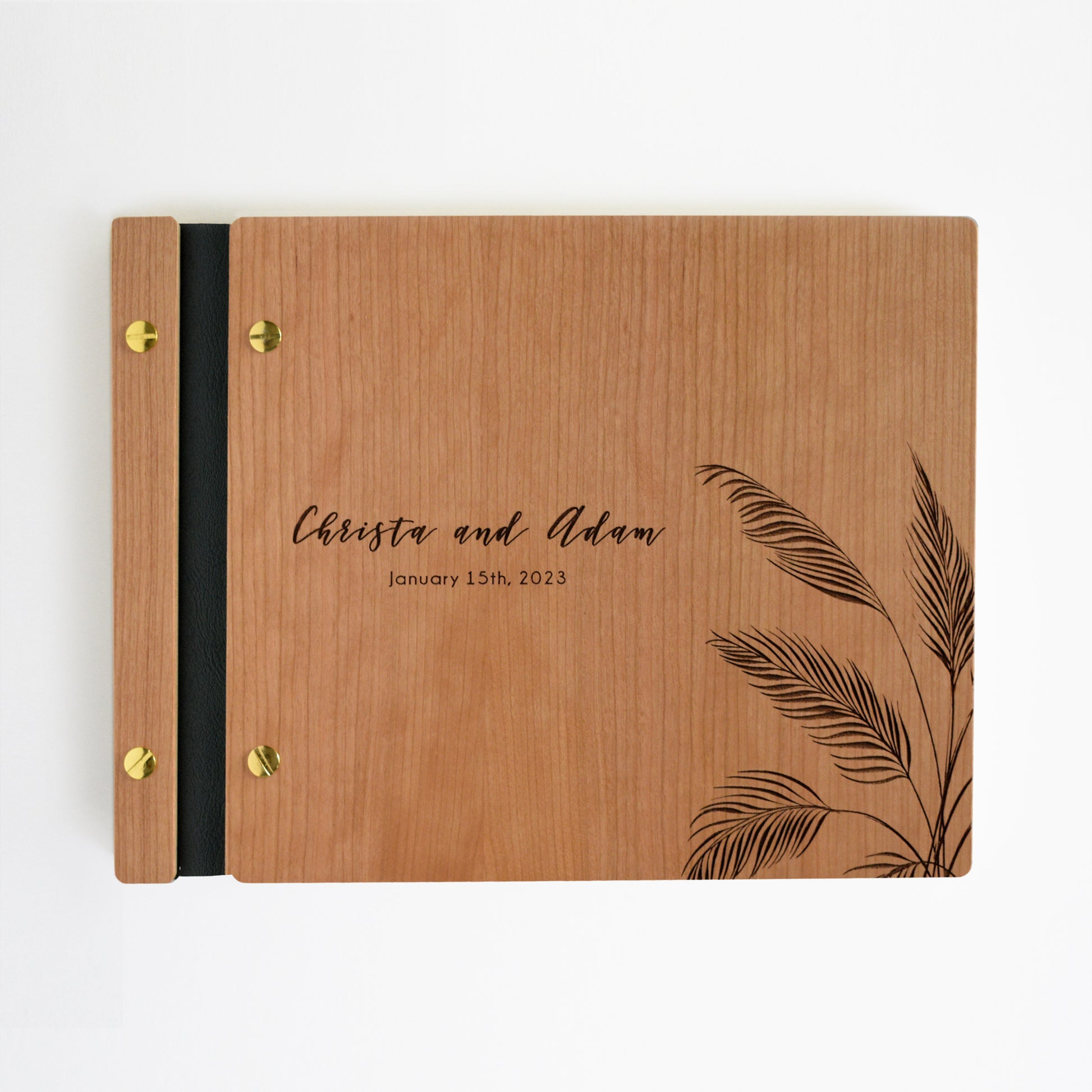 An 8.5x11" guest book lies on a table. Made of cherry wood, black vegan leather binding, and copper hardware. The front cover reads “Christina and Adam, January 15th, 2023.” 
