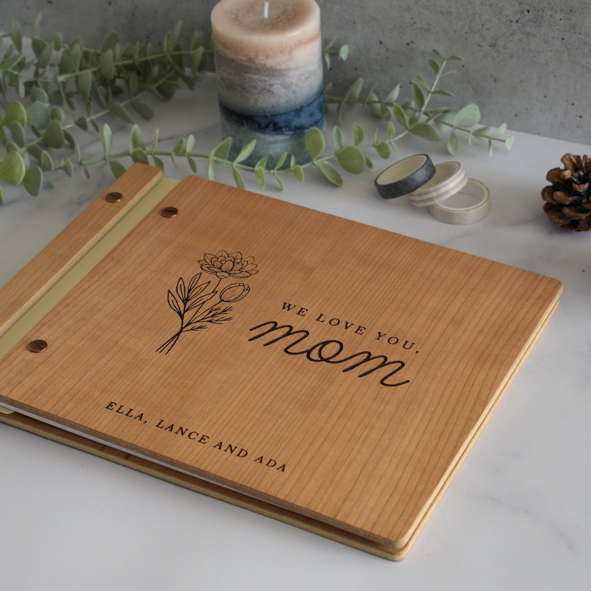 An 8.5x11" sized guest book lies on a table with greenery and a candle. This custom photo album includes a personalized engraving that reads We Love You, Mom with flowers.