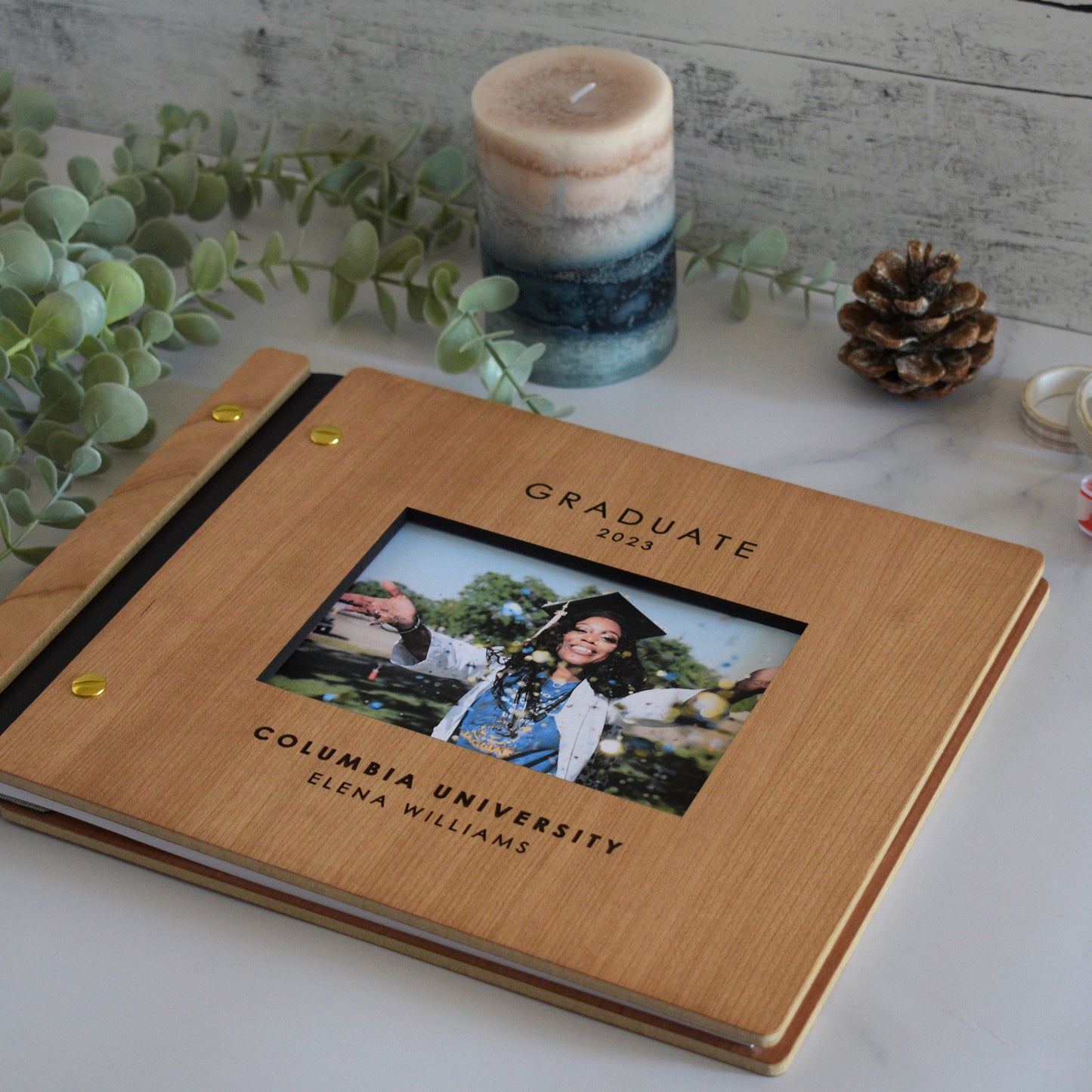 An 8.5x11" sized guest book lies on a table with greenery and a candle. The guest book includes a personalized engraving that reads Graduate 2023 along with the University name and Student name. The guest book also includes a cut out frame with a photo of the graduate.