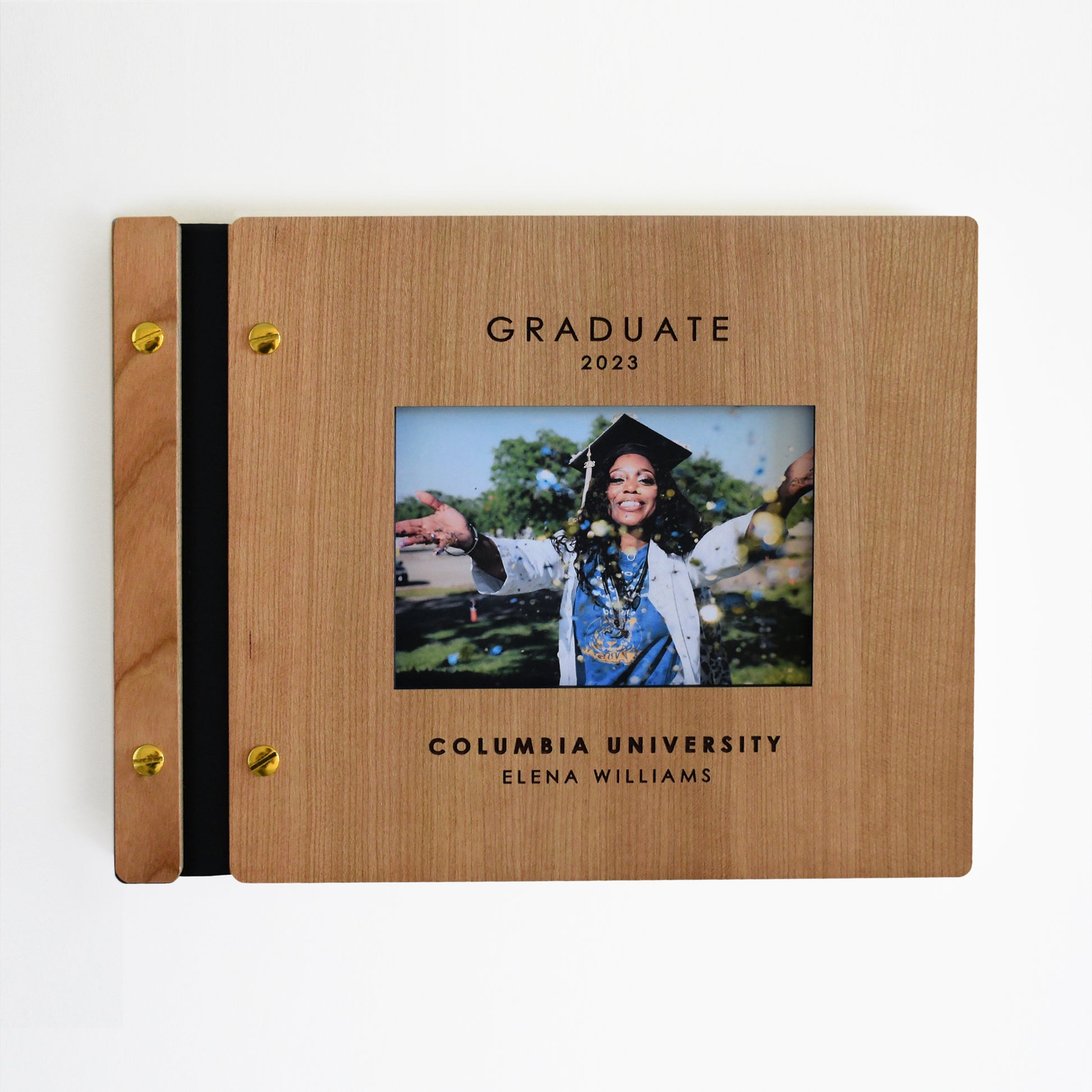 An 8.5x11" sized guest book stands on a table with a white background. The graduation guest book is made from cherry wood, black vegan leather, and gold hardware. This is the perfect addition to a photo booth or polaroid table for guests to sign and post polaroid photos in!