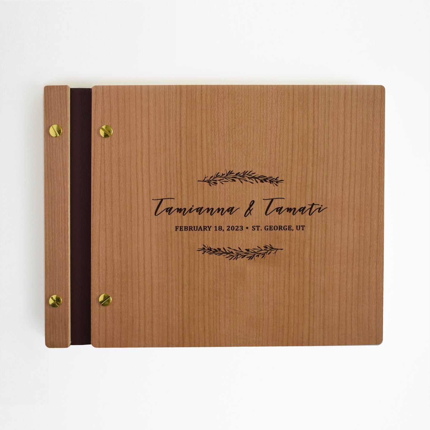 An 8.5x11" guest book lies on a table. Made of cherry wood, black vegan leather binding, and copper hardware. The front cover includes an engraved design with personalized names. 
